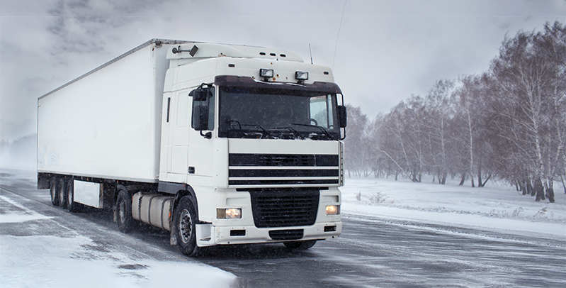 Large commercial truck driving on a snow-covered road during winter, with reduced visibility and icy conditions.