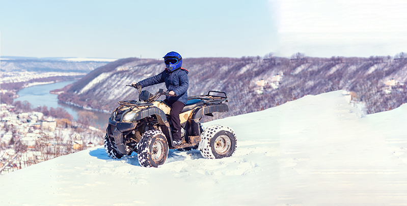 Person riding an ATV across a snowy mountain landscape, wearing winter gear and helmet in cold outdoor conditions.